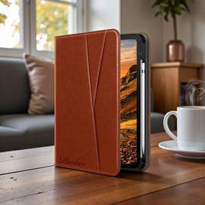May include: A brown leather tablet case with a stylus. The case has a stitched design and the name "Parker" embossed on the front. The tablet displays a landscape image. A white coffee cup sits on a saucer on a wooden table.