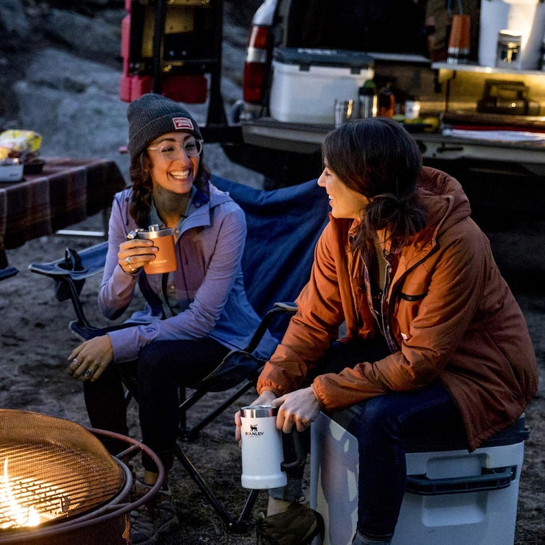 May include: Two people are seated outdoors, each holding a Stanley mug. One mug is orange, the other is white with the Stanley logo. They are wearing jackets and a beanie. The scene suggests a camping or outdoor activity.