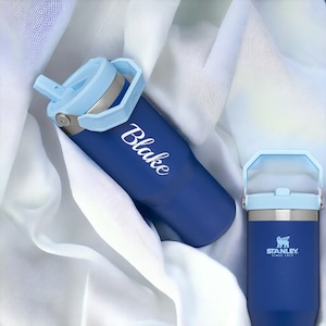 May include: Two blue stainless steel water bottles with a light blue lid and handle. The bottle on the left has the name "Blake" written on it in white. The bottle on the right has the Stanley logo and "Since 1913" printed on it.