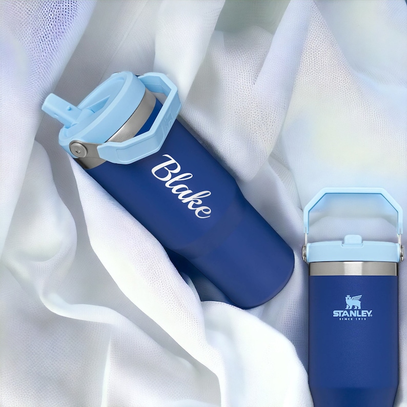 May include: Two blue stainless steel water bottles with a light blue lid and handle. The bottle on the left has the name "Blake" written on it in white. The bottle on the right has the Stanley logo and "Since 1913" printed on it.