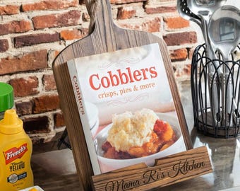 May include: Wooden cookbook stand with a book titled "Cobblers" displayed. The book cover features a photo of a cobbler dessert. The stand is engraved with "Mama Ri's Kitchen". A bottle of yellow mustard and a small dish of cheese cubes are in the background.