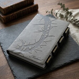 May include: Gray leather journal with a debossed tree design and the name "Michael" on the cover. The journal has black tabbed dividers labeled with book names. The journal is on a black slate surface.