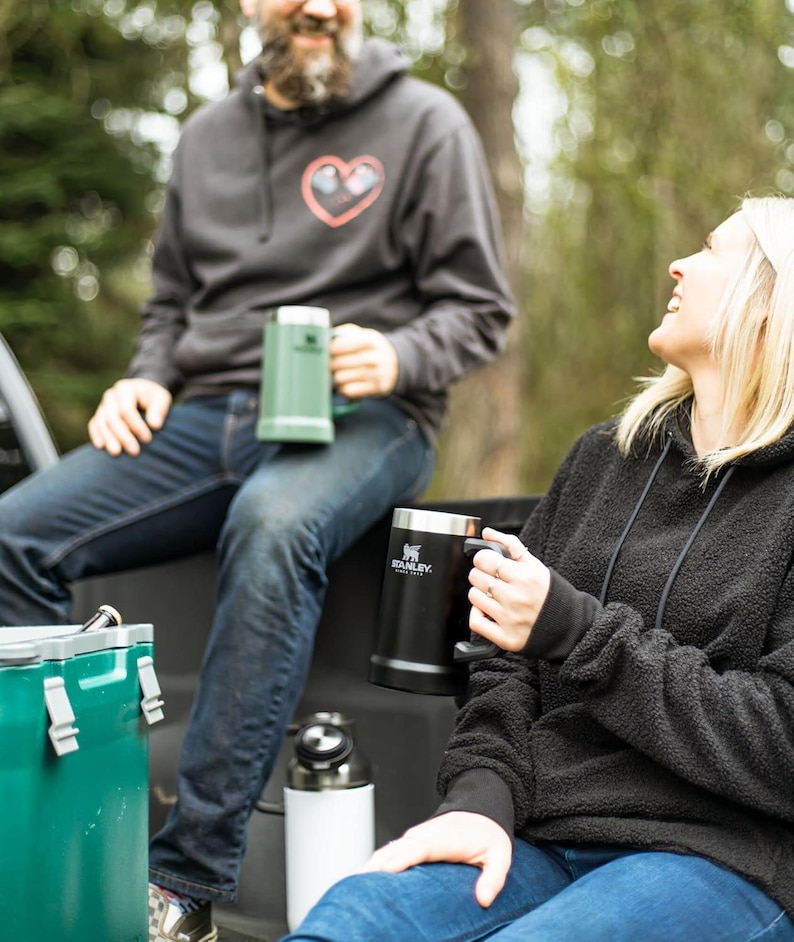 May include: Two people are sitting outdoors, each holding a Stanley mug. One mug is black with a silver rim, the other is green. The person in the background wears a grey hoodie with a heart design. The other person wears a black hoodie.