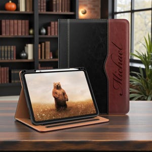 May include: A brown leather tablet case with a black accent and the name "Michael" in cursive. The tablet displays a picture of a bear standing in a field. The case is open, and a stylus is visible.