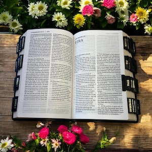 Peut inclure: Bible ouverte avec des onglets noirs indiquant les livres, dont &laquo; Ezra &raquo; et &laquo; N&eacute;h&eacute;mie &raquo;. Les pages sont ouvertes sur le texte, entour&eacute;es de fleurs roses, blanches et jaunes, sur une surface en bois.