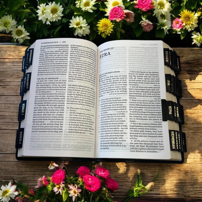 Peut inclure: Bible ouverte avec des onglets noirs indiquant les livres, dont &laquo; Ezra &raquo; et &laquo; N&eacute;h&eacute;mie &raquo;. Les pages sont ouvertes sur le texte, entour&eacute;es de fleurs roses, blanches et jaunes, sur une surface en bois.