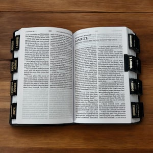 K&ouml;nnte beinhalten: Ge&ouml;ffnete Bibel mit schwarzen Registerkarten, die mit Buchnamen wie Genesis, Exodus und Samuel beschriftet sind. Die Seiten sind zum Text ge&ouml;ffnet, mit Fokus auf den Text und den Buchtitel.