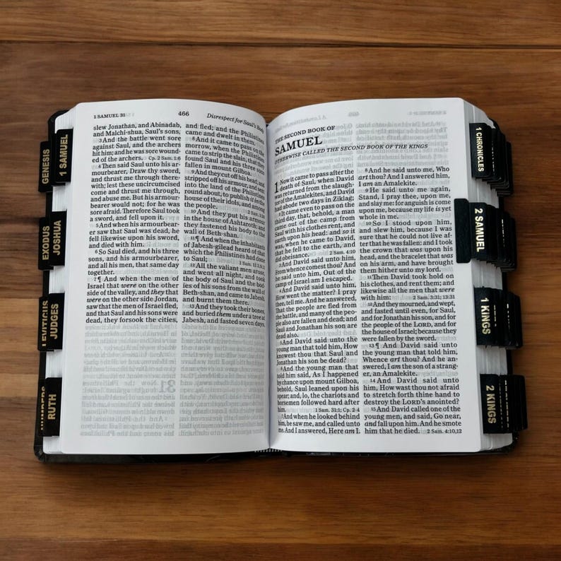 K&ouml;nnte beinhalten: Ge&ouml;ffnete Bibel mit schwarzen Registerkarten, die mit Buchnamen wie Genesis, Exodus und Samuel beschriftet sind. Die Seiten sind zum Text ge&ouml;ffnet, mit Fokus auf den Text und den Buchtitel.