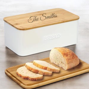 May include: White metal bread box with a bamboo lid. The lid has the text "The Smiths" engraved on it. The front of the box has the word "BREAD" embossed on it. A loaf of bread and slices of bread are on a bamboo cutting board.