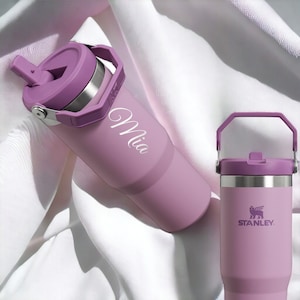 May include: Two purple stainless steel water bottles with lids. One bottle has a carrying handle and the name "Mia" engraved on the side. The other bottle has the Stanley logo on the side.