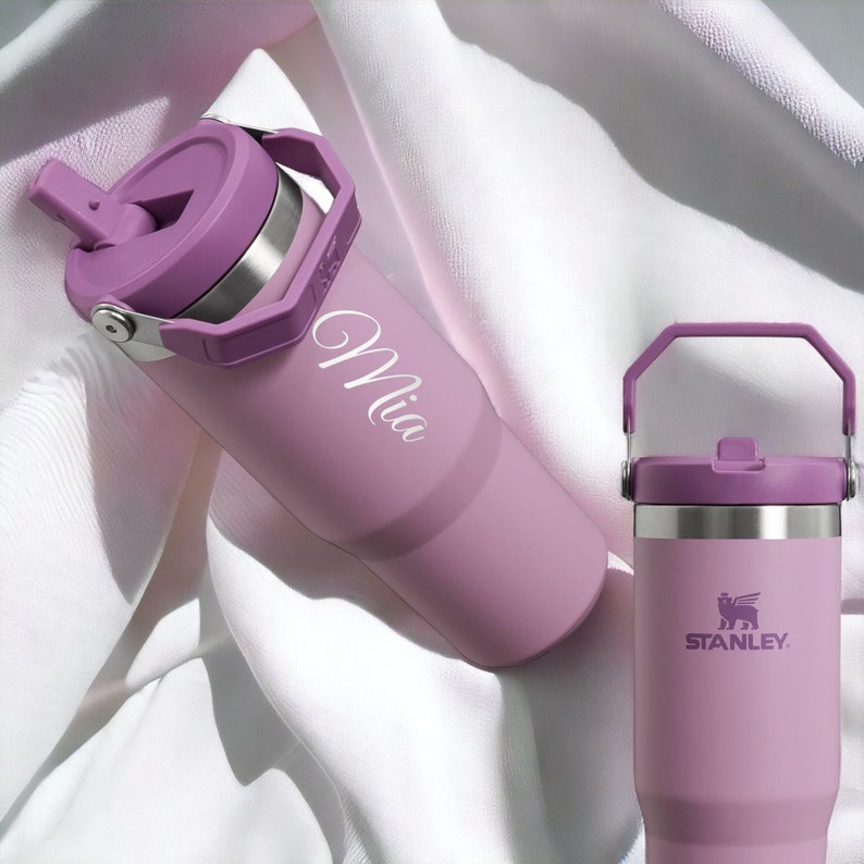 May include: Two purple stainless steel water bottles with lids. One bottle has a carrying handle and the name "Mia" engraved on the side. The other bottle has the Stanley logo on the side.