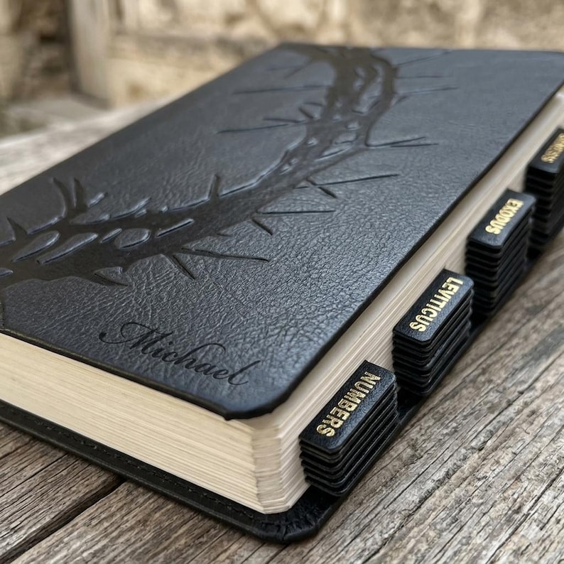 K&ouml;nnte beinhalten: Schwarz ledergebundenes Buch mit goldfarbenen Seiten und gepr&auml;gtem Dornendesign. Mit dem Namen "Michael" und schwarzen Laschen mit der Aufschrift "Numbers", "Leviticus" und "Exodus". Das Buch ist ge&ouml;ffnet und zeigt die Seiten.