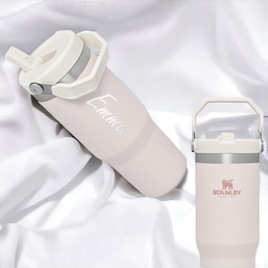 May include: Two pastel pink stainless steel water bottles with white lids and handles. The bottle on the left has the name "Emma" written on it. The bottle on the right has the Stanley logo and the text "Since 1913".