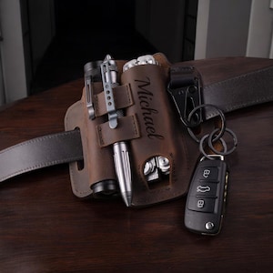 Pode incluir: Um cinto de couro castanho com uma bolsa de couro personalizada para transportar ferramentas e um chaveiro. A bolsa tem o nome "Michael" gravado.