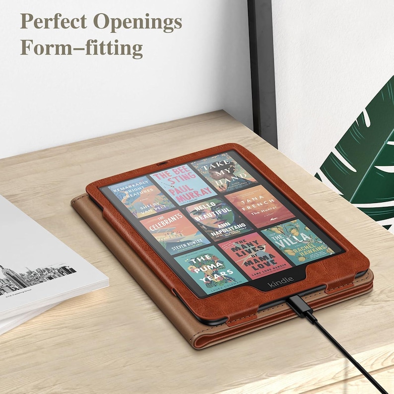 May include: A brown leather-textured Kindle e-reader case with a form-fitting design and precise cutouts. The device screen displays a grid of nine book covers, and a black charging cable is plugged into the bottom port of the e-reader.