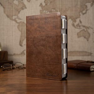 May include: A brown leather Bible with a personalized name, featuring labeled tabs for easy navigation. The cover has a textured appearance. The Bible is open, revealing the pages and tabs. The background includes a world map.