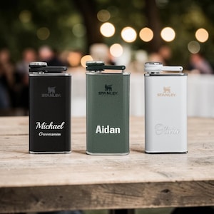 May include: Three personalised Stanley hip flasks in black, green, and white, each with a name and the Stanley logo. The black flask says "Michael Groomsman", the green flask says "Aidan", and the white flask says "Olivia". They are on a wooden table.