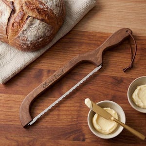 May include: A wooden bread bow knife with a serrated metal blade and a leather hanging loop. The wooden handle is engraved with the text Olivia's Bread. The knife is positioned near a loaf of bread and a small bowl containing a scoop of butter.