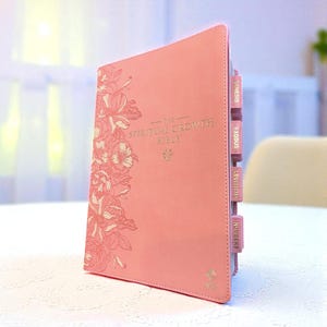 May include: A pink Bible with the text "The Spiritual Growth Bible" in silver lettering. The cover features a floral design on the left side. The book has pink tabs with the names of the books of the Bible.