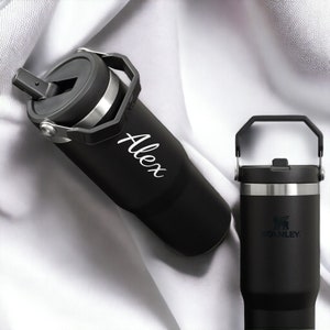May include: Two black stainless steel travel mugs with a carrying handle. The mug on the left has the name "Alex" written on it. The mug on the right has the Stanley logo.
