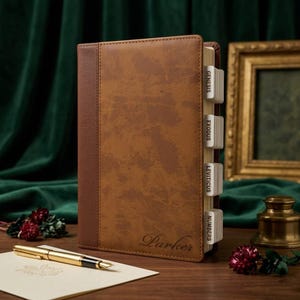 May include: A brown leather-bound book with a textured cover and a dark brown spine. The book has labeled tabs and the name "Parker" is embossed on the cover. A gold pen and a card are in the foreground.
