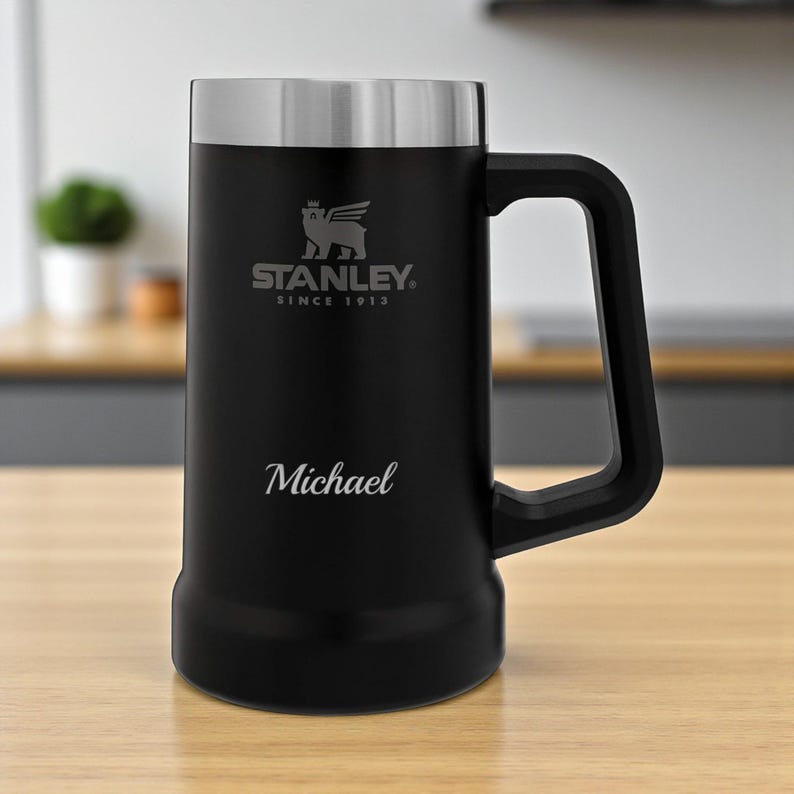 May include: Black Stanley beer stein with a silver lid and handle. The stein features the Stanley logo and the name "Michael" engraved on the front. The stein is on a wooden surface.