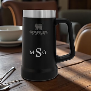May include: Black Stanley beer stein with a silver lid and handle. The stein features the Stanley logo and the text "SINCE 1913". The initials "MSG" are engraved on the front. The stein is on a wooden table.