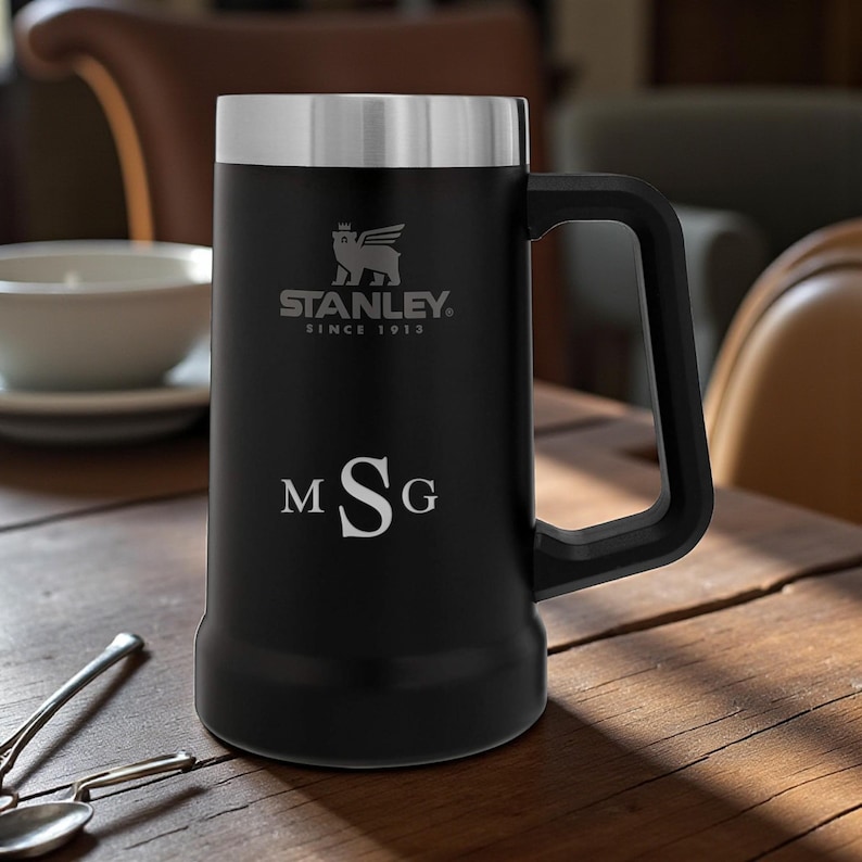 May include: Black Stanley beer stein with a silver lid and handle. The stein features the Stanley logo and the text "SINCE 1913". The initials "MSG" are engraved on the front. The stein is on a wooden table.