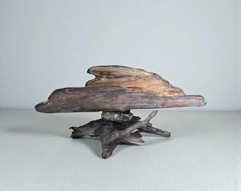 May include: A natural driftwood sculpture. The piece features a large, horizontal piece of driftwood balanced on a smaller, root-like base. The wood has a mix of brown and tan tones, with a rough, textured surface. The sculpture is a unique decorative item.