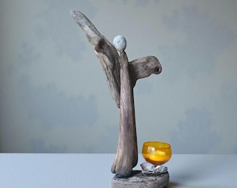 May include: A driftwood sculpture with a small, round stone at the top. The sculpture has a natural, weathered appearance. An orange glass votive holder sits on a stone base, adding a pop of color.