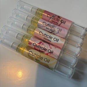 May include: Five cuticle oil pens are displayed. Each pen features a clear plastic tube, a white applicator, and a label with the words "Cuticle Oil" and a unique name: Serenity, Noir Op, Paradise, Rouge, and Uplift. The oil inside is yellow.