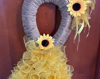 May include: A burlap wreath with two yellow sunflowers and a yellow mesh bow. The wreath is hanging on a wooden door.