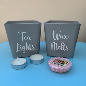 May include: Two grey square containers with white lettering. The container on the left says "Tea Lights" and the container on the right says "Wax Melts". There are two white tea lights and a pink wax melt on a blue surface.