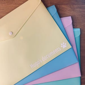 May include: A collection of pastel document wallets in yellow, blue, pink, and teal. The yellow wallet has a white press stud closure. The text "Hugo's documents" and a paw print are printed on the front of the yellow wallet.
