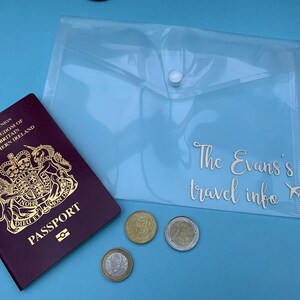 May include: A clear plastic travel document holder with a white button closure. The holder has the text "The Evans's travel info" printed on it. A British passport, a few coins, and a small airplane icon are visible in the background.