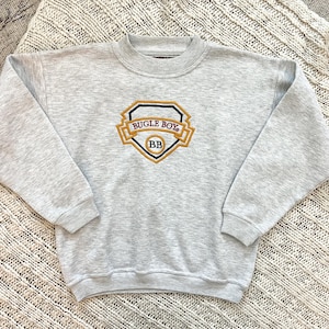 May include: Light gray sweatshirt with a crew neck and long sleeves. The front features an embroidered emblem with the words "BUGLE BOY" and "BB" in gold and navy blue.