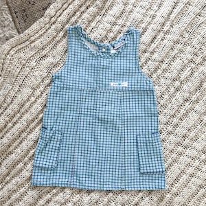 May include: A sleeveless jumper dress in a blue and white checkered pattern featuring a round neckline, two front patch pockets, and a small white rectangular label on the chest with the text "Bright Day" printed in blue letters.