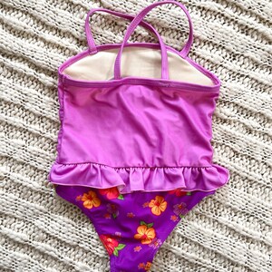 Puede incluir: Un ba&ntilde;ador de una pieza en tonos morados y rosas, con un estampado floral en la parte inferior. El ba&ntilde;ador tiene un ribete con volantes y tirantes finos. El fondo es una superficie texturizada blanquecina.