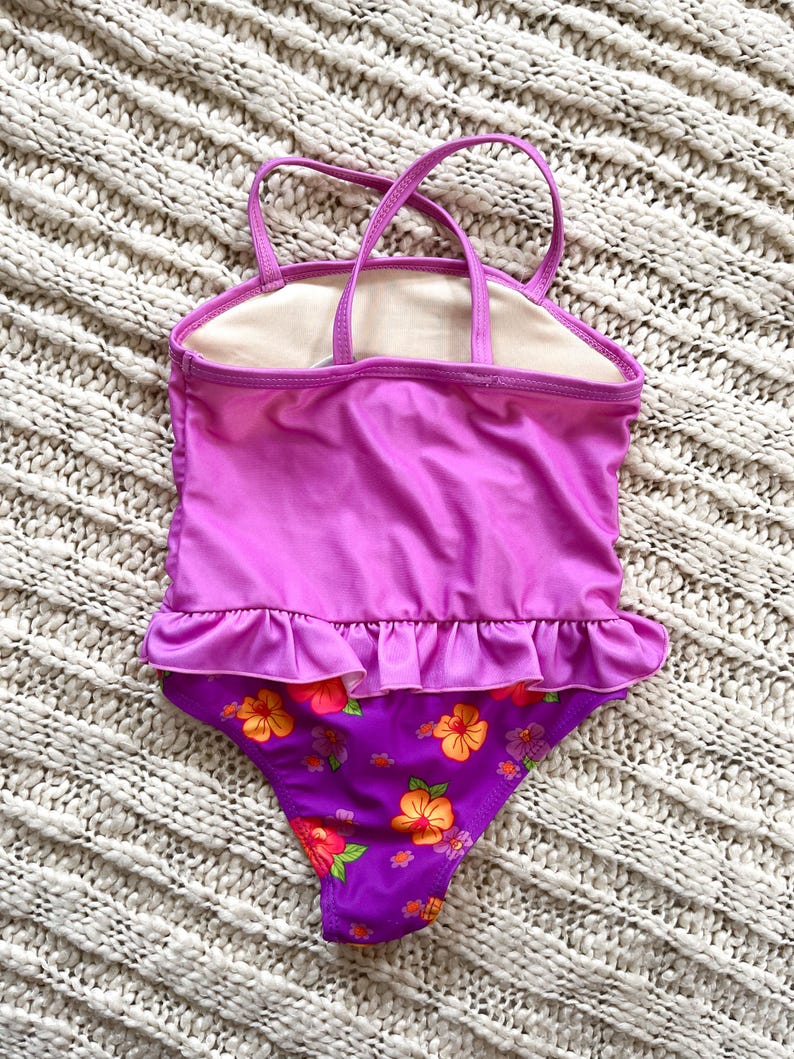 Puede incluir: Un ba&ntilde;ador de una pieza en tonos morados y rosas, con un estampado floral en la parte inferior. El ba&ntilde;ador tiene un ribete con volantes y tirantes finos. El fondo es una superficie texturizada blanquecina.