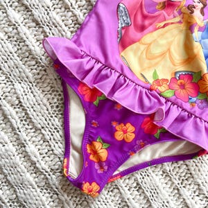 Puede incluir: Traje de ba&ntilde;o morado con ribete de volantes y estampado floral. El traje de ba&ntilde;o presenta un gr&aacute;fico de princesas animadas. La parte inferior del traje de ba&ntilde;o tiene un estampado floral con flores naranjas y rosas.