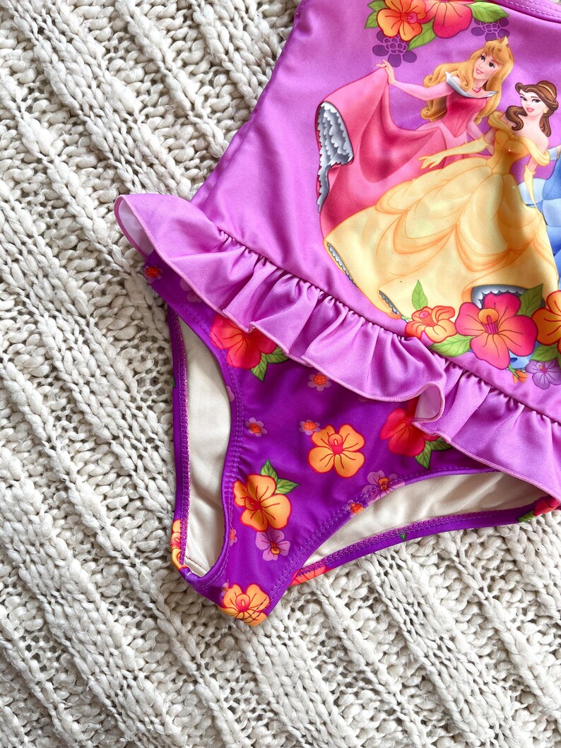 Puede incluir: Traje de ba&ntilde;o morado con ribete de volantes y estampado floral. El traje de ba&ntilde;o presenta un gr&aacute;fico de princesas animadas. La parte inferior del traje de ba&ntilde;o tiene un estampado floral con flores naranjas y rosas.
