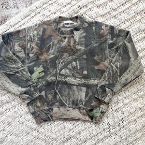 May include: A long-sleeve crewneck sweatshirt featuring a realistic forest camouflage pattern in shades of brown, green, gray, and black. The garment has a ribbed collar, cuffs, and hem, with a small rectangular white brand label at the neckline.