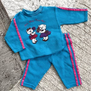 May include: A blue fleece baby tracksuit with pink stripes. The top features a graphic of two teddy bears with the text "It's Party Time Bear".