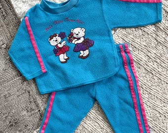 Puede incluir: Un chándal de bebé de franela azul con rayas rosas. La parte superior presenta un gráfico de dos ositos de peluche con el texto "It's Party Time Bear".