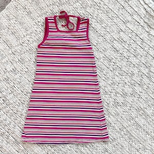 May include: A striped sleeveless dress in pink, purple, and white. The dress features a halter neck and a simple, straight cut. The garment is displayed on a textured, off-white surface.