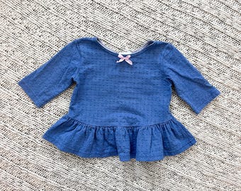 May include: A blue top with three-quarter sleeves and a peplum hem. The top features a textured dot pattern and a small pink bow at the neckline. The garment is displayed on a textured, off-white surface.