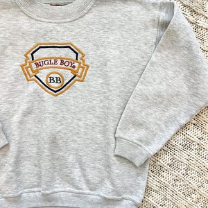 May include: A light gray sweatshirt with a crew neck and long sleeves. The front features an embroidered emblem with the words "BUGLE BOY" and "BB" in a gold and navy blue design. The sweatshirt is made of a soft material.