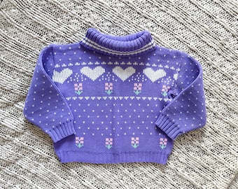 Puede incluir: Un suéter morado de cuello alto con un estampado floral y de corazón blanco y rosa. El suéter tiene cuello y puños acanalados, y pequeños puntos blancos están dispersos por la tela. El suéter está hecho de un material de punto suave.
