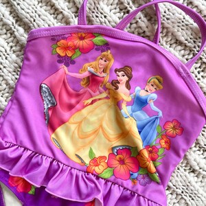 Puede incluir: Traje de ba&ntilde;o de una pieza morado con un gr&aacute;fico de tres princesas de Disney con sus vestidos. El traje de ba&ntilde;o tiene un ribete con volantes y correas. Acentos florales en naranja, rosa y amarillo decoran el traje de ba&ntilde;o. La parte inferior es morada con motivos florales.