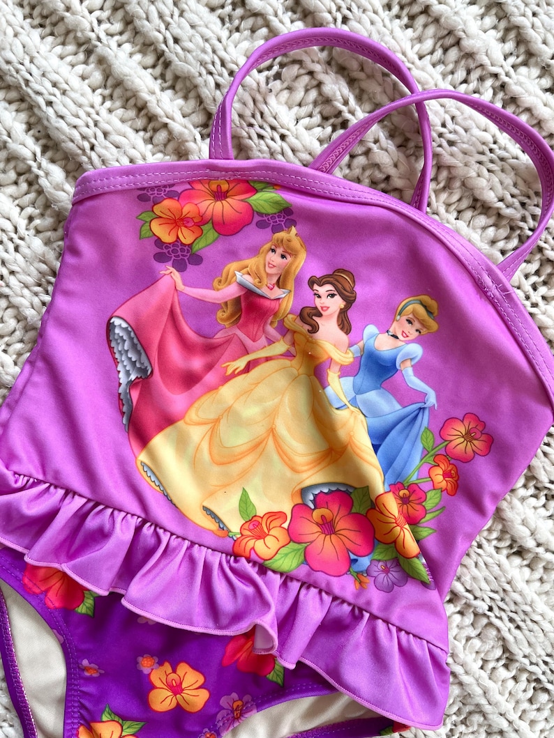 Puede incluir: Traje de ba&ntilde;o de una pieza morado con un gr&aacute;fico de tres princesas de Disney con sus vestidos. El traje de ba&ntilde;o tiene un ribete con volantes y correas. Acentos florales en naranja, rosa y amarillo decoran el traje de ba&ntilde;o. La parte inferior es morada con motivos florales.